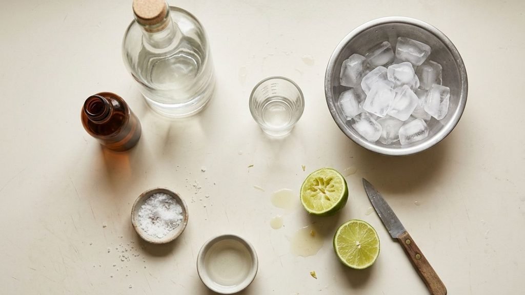 Margarita ingredients arranged on a stone surface, including limes, tequila, orange liqueur, syrup, salt and ice.