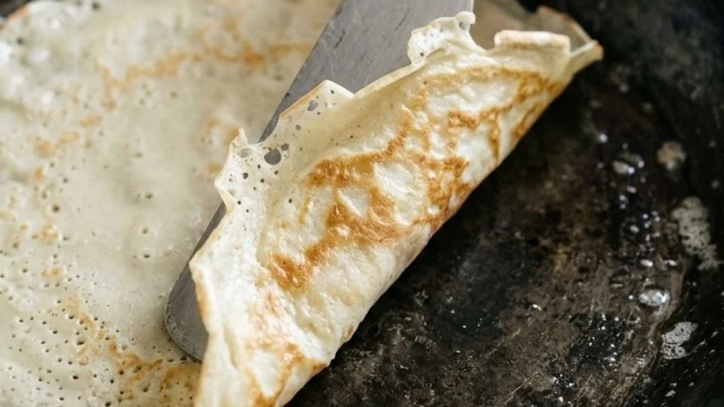 A metal spatula lifts the edge of a lightly browned thin pancake in a frying pan.