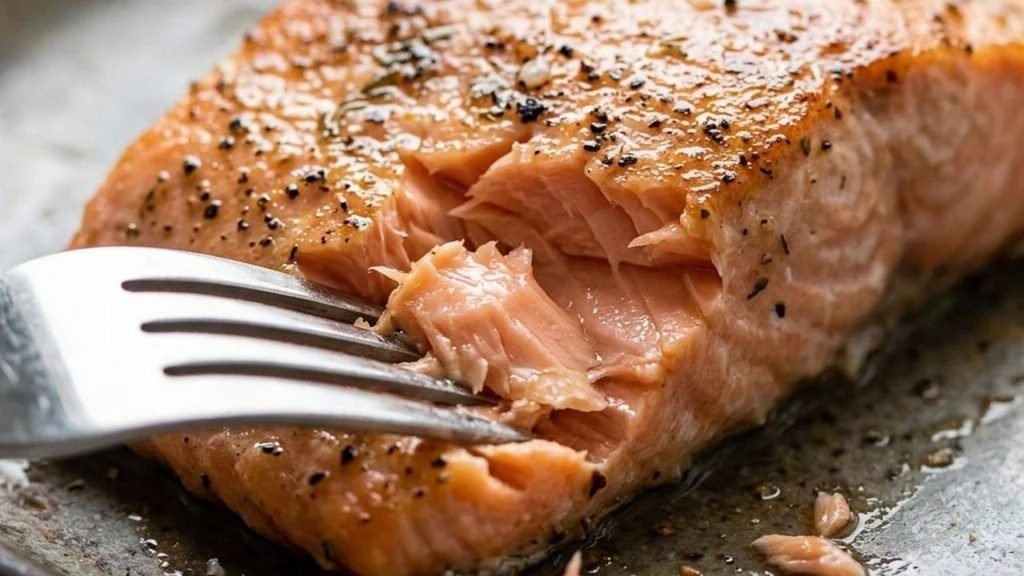 A fork separates the moist pink flakes of baked salmon beneath its seasoned surface.