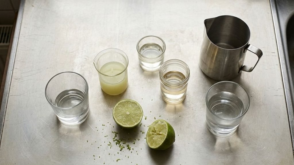 Measured margarita ingredients in clear glasses beside cut limes and a stainless-steel measuring jug.